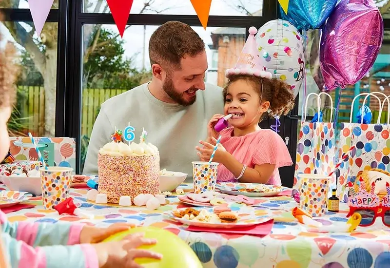 A colorful birthday party scene with a table covered in a polka-dot tablecloth, matching cups and plates, a large cake decorated with sprinkles and a number 6 candle, wrapped gifts, balloons, and festive banners in the background.